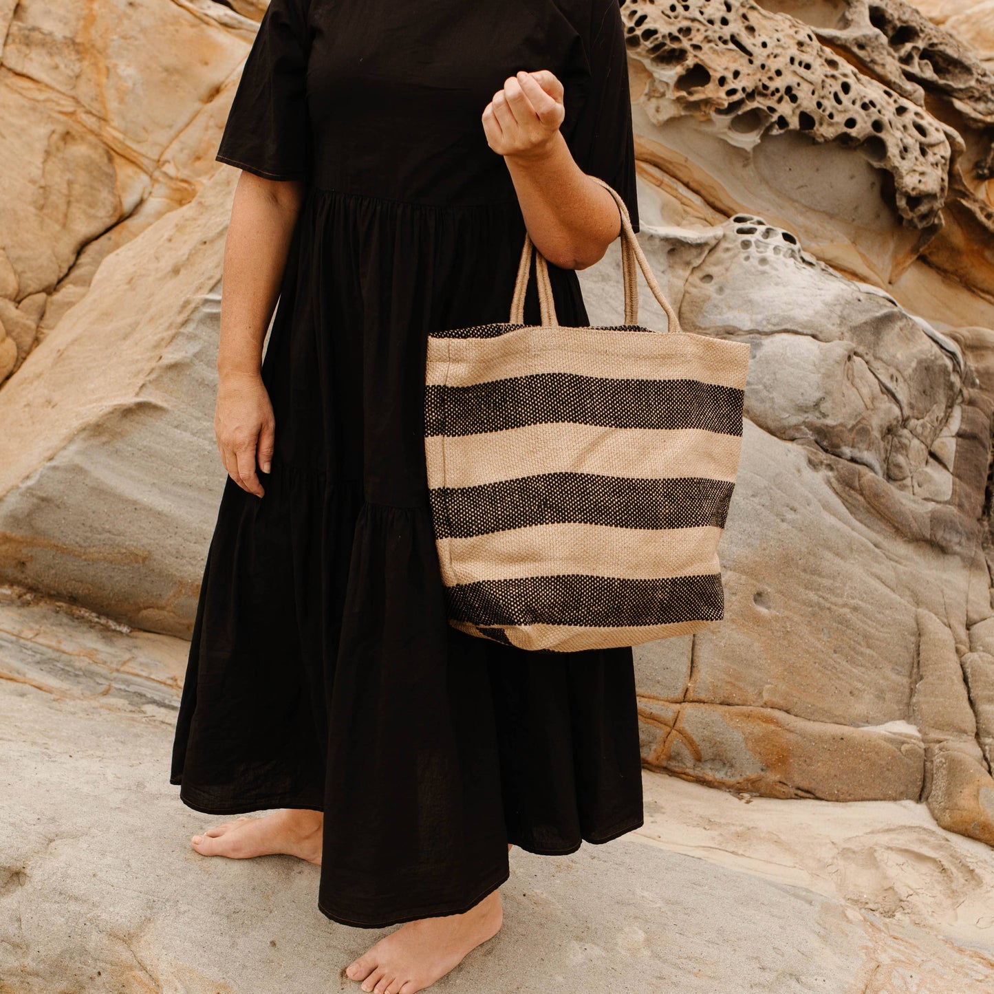 Woman in a black dress holding a striped bag on a rocky beach.