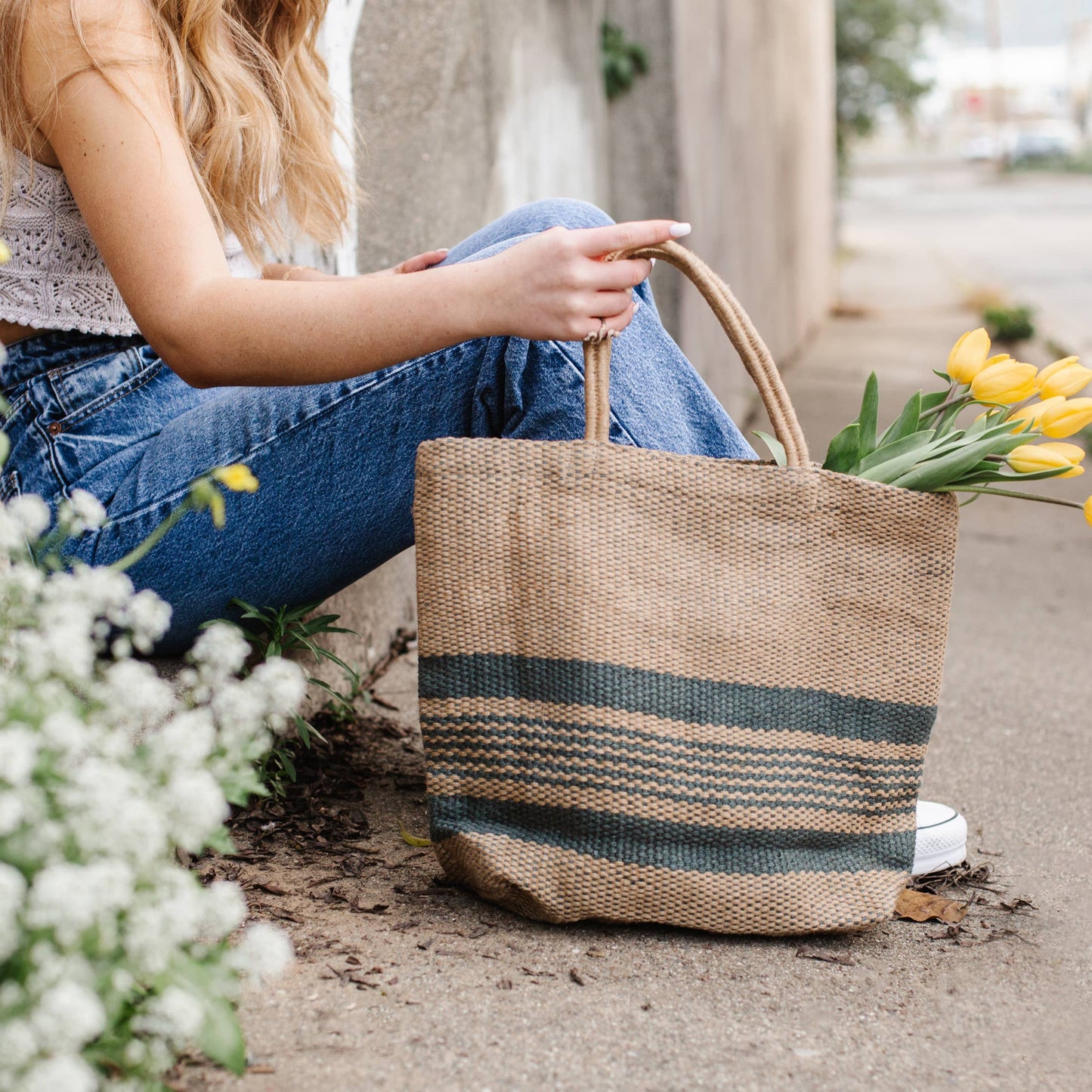 Person holding a woven bag with flowers, sitting on a concrete surface with plants around.