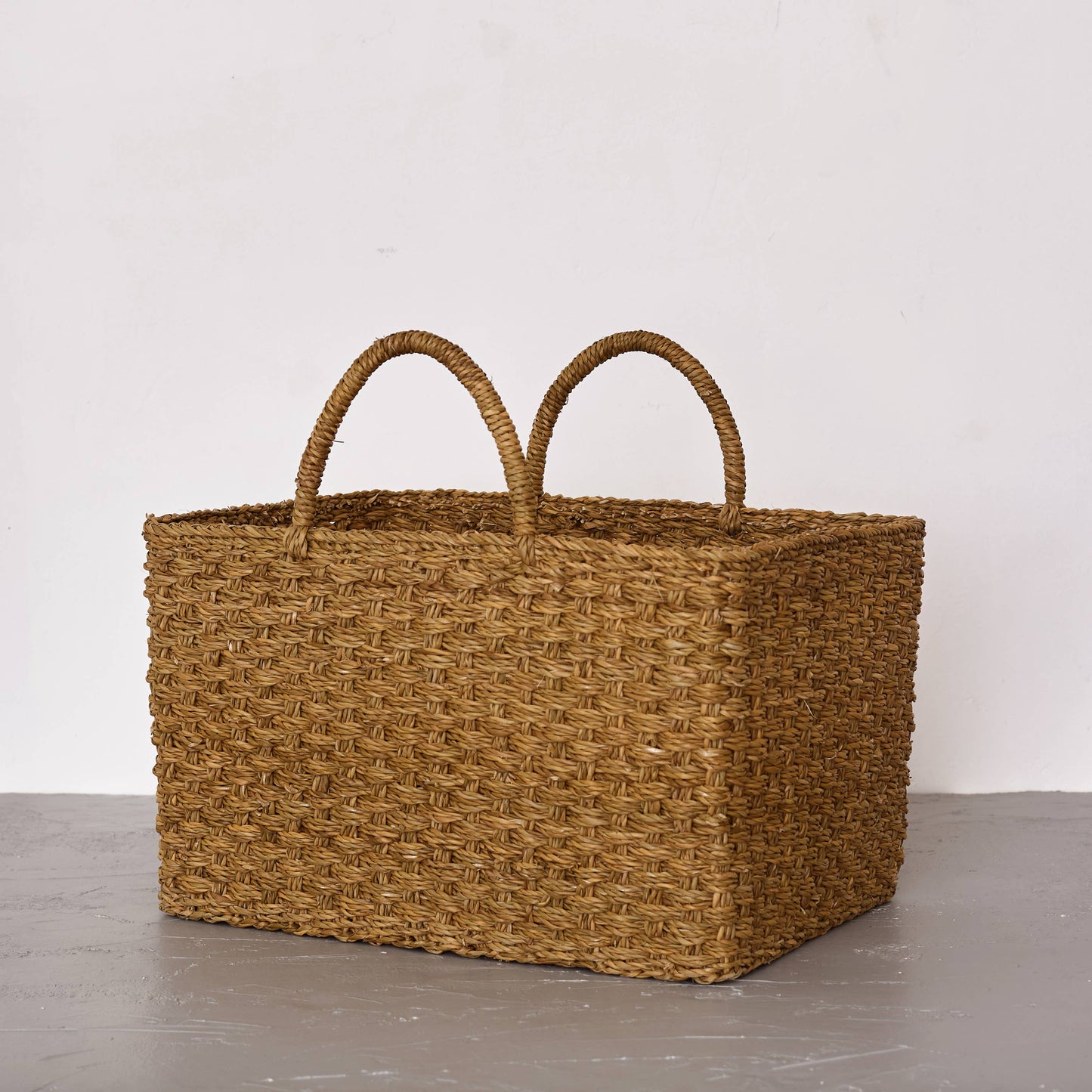 Woven brown basket with handles on a gray surface and white background