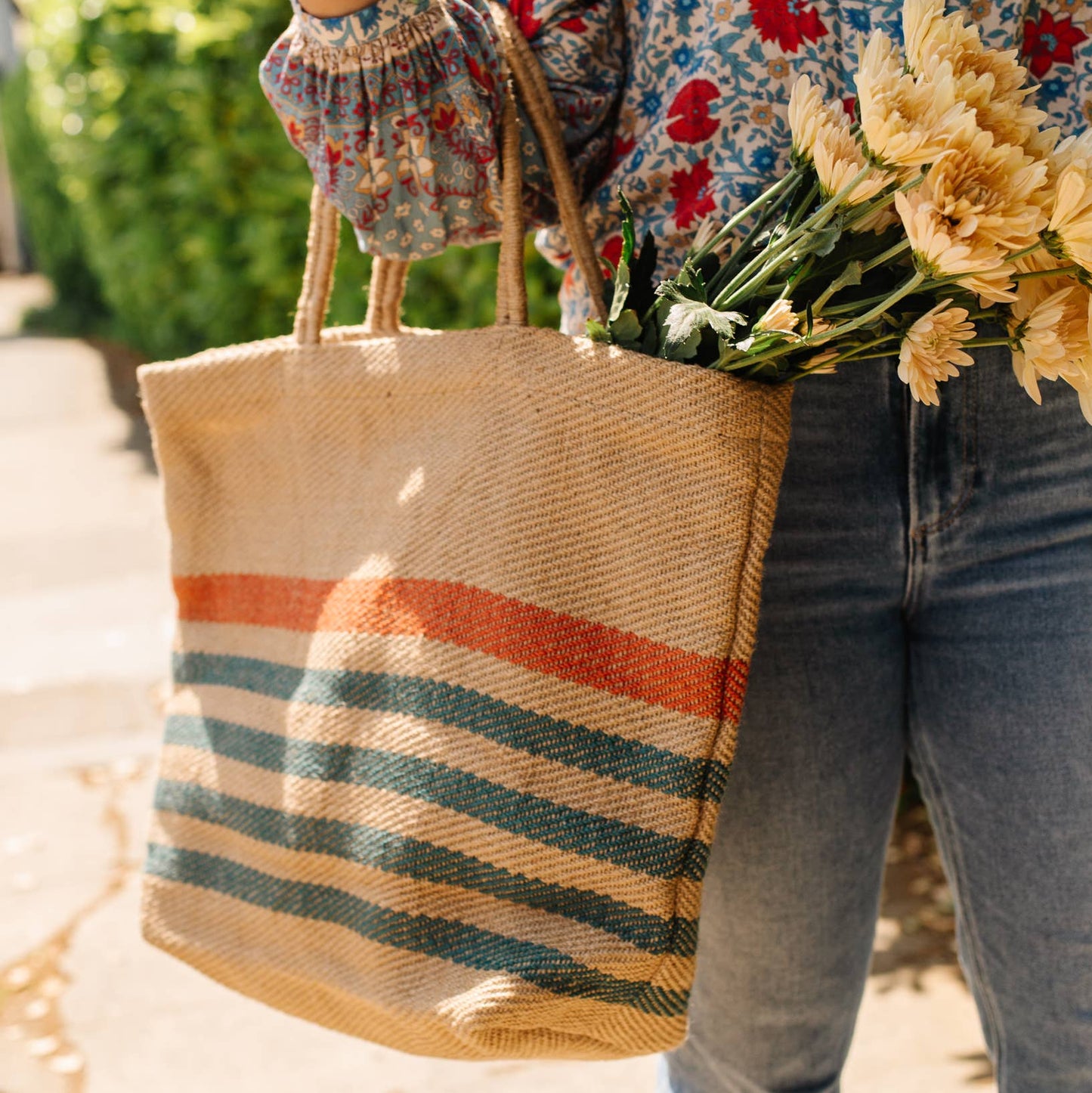 Person holding a striped tote bag and flowers, wearing a floral shirt and jeans.