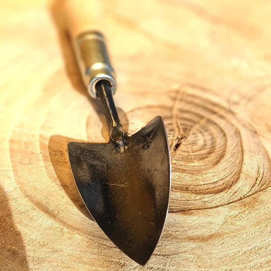 A small hand trowel with a dark metal blade and a light-colored wooden handle, resting on a textured wood surface.