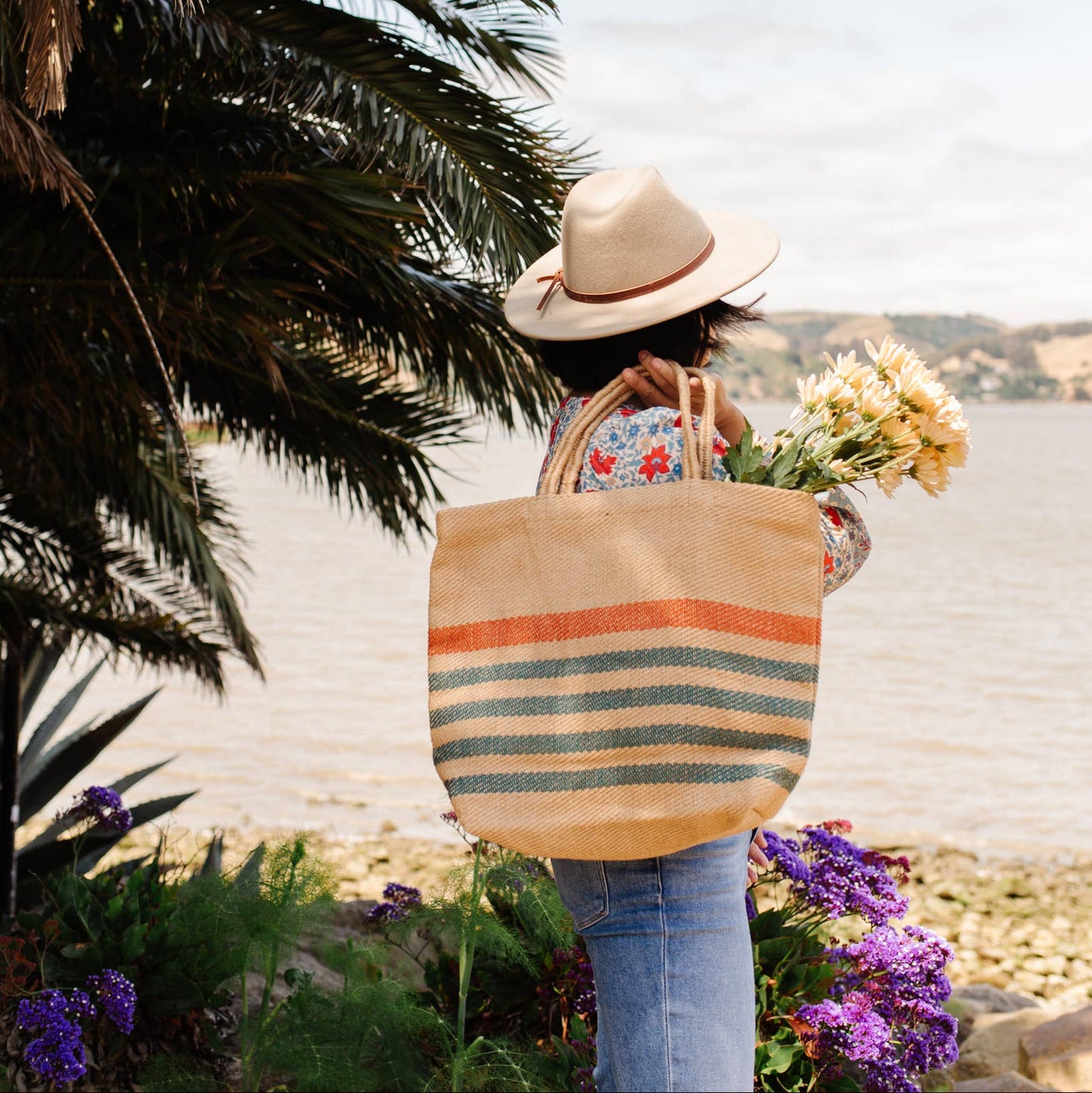 Person with a straw hat and floral bag by a palm tree on a beach