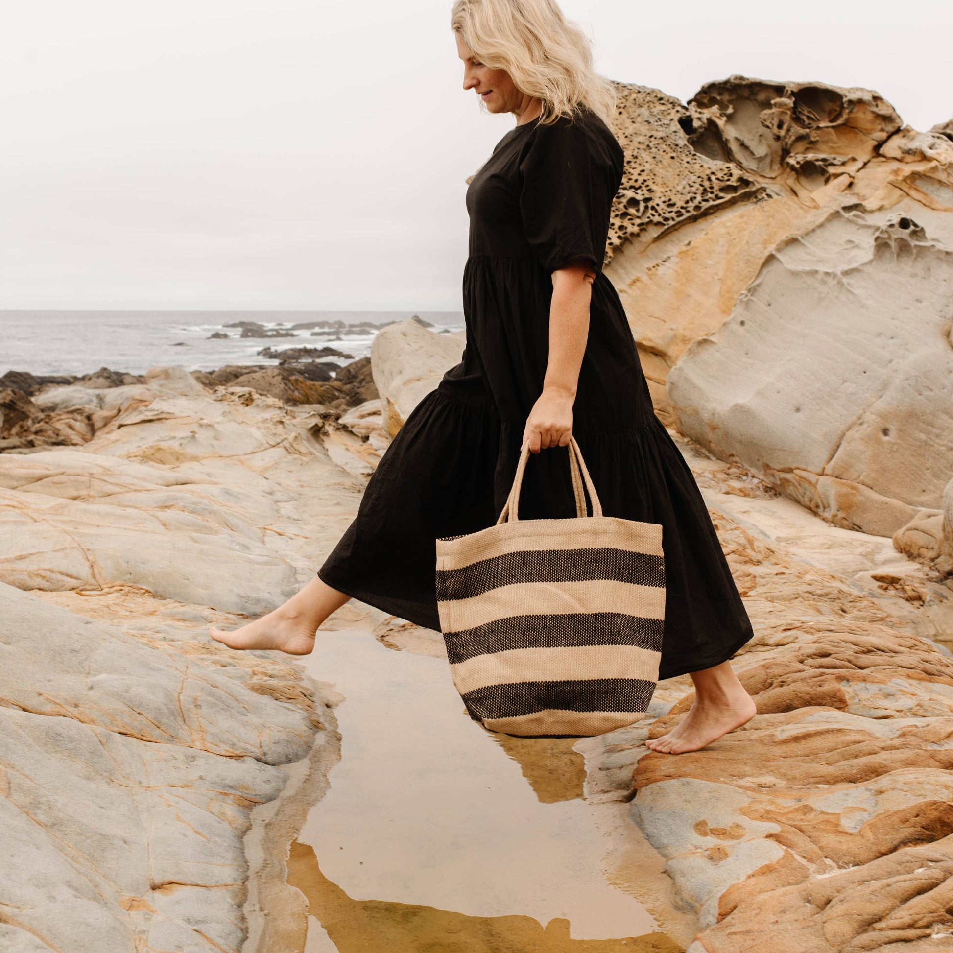 Woman in a black dress holding a striped bag on a rocky beach.